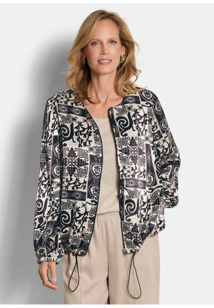 Femme portant une veste zippée à motifs noir et blanc par-dessus un haut beige clair et un pantalon beige, debout avec une main dans la poche.