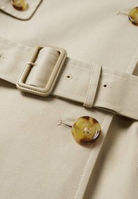 Béžová tkanina s širokým opaskem a obdélníkovou sponou. Dva knoflíky z mramorovaného žluto-hnědého materiálu. Obsahuje vyšívané detaily a malé dírky pro knoflíky.