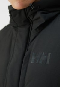 Jachetă neagră cu glugă și guler înalt, cu un logo mat "HH" pe piept, realizată dintr-un material durabil cu o textură netedă.
