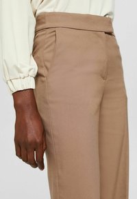 Gros plan d'une personne portant un pantalon beige taille haute et un chemisier crème à manches longues avec poignets élastiques, debout avec un bras détendu.