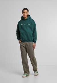 Sudadera verde con un bolsillo frontal, con texto impreso en blanco, combinada con pantalones sueltos de color gris claro y zapatillas de colores.