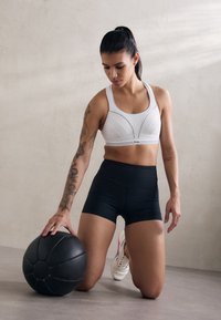Sports bra in white with structured panels, paired with black high-waisted shorts. A black medicine ball is positioned beside a kneeling model.