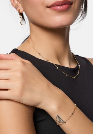 Colliers superposés en ton or avec des accents en barre et bracelet assorti. La texture est lisse ; les boucles d'oreilles présentent un design en anneau avec des éléments pendants.