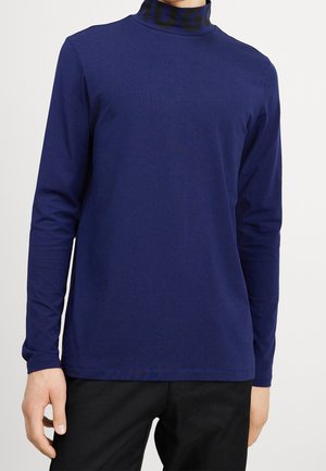Marineblaues Langarmshirt mit hohem Kragen und schwarzem Logoband am Hals. Glatter Stoff mit figurbetonter Passform, minimale Nähte.