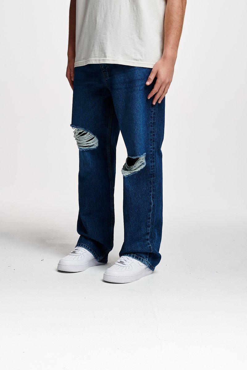 Jeans de ganga azuis com remendos em torno dos joelhos, corte relaxado e bordas desfiadas; acompanhados de ténis brancos.