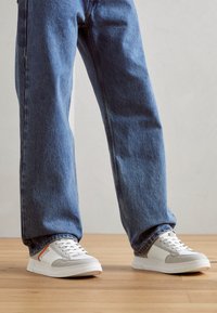 Sneakers bianchi e grigi con una parte superiore in pelle liscia, dettagli in tessuto e cuciture a vista, abbinati a jeans in denim blu dalla vestibilità rilassata.