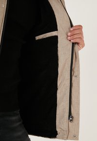 Bežs žakets ar teksturētu virsmu, ar melnu polāru odere, ar rāvējslēdzēja aizdari un mazu iekšējo kabatu gar šuvi.