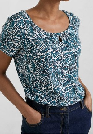 Vrouw draagt een blauw-wit topje met korte mouwen en een keyhole-halslijn met een bladmotive, ingestopt in donkerblauwe denim jeans.