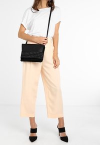 Černá křížová taška s překlapávacím designem, zdobená stříbrnými cvočky, vyrobená z texturovaného materiálu. Spojena s širokými kalhotami v jemné broskvové barvě.