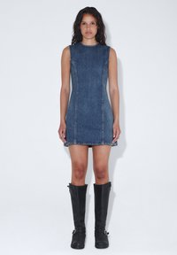 Sininen denimpitkä hihaton mekko korkealla kaula-aukolla, kontrastivärillä ommeltu, hieman levenevä helma, yhdistettynä polven korkuisiin mustiin saappaisiin.