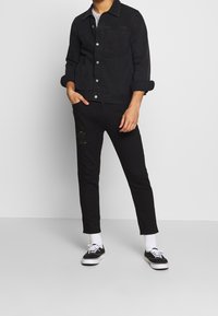 Svart denimjacka med knappstängning och en enda bröstficka, matchad med svarta smala jeans med slitage. Svarta sneakers.