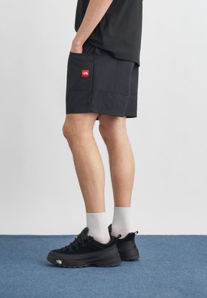 Persona con pantalones cortos negros con logo rojo, zapatillas negras con logo blanco, calcetines blancos, de pie sobre una alfombra azul contra una pared lisa.