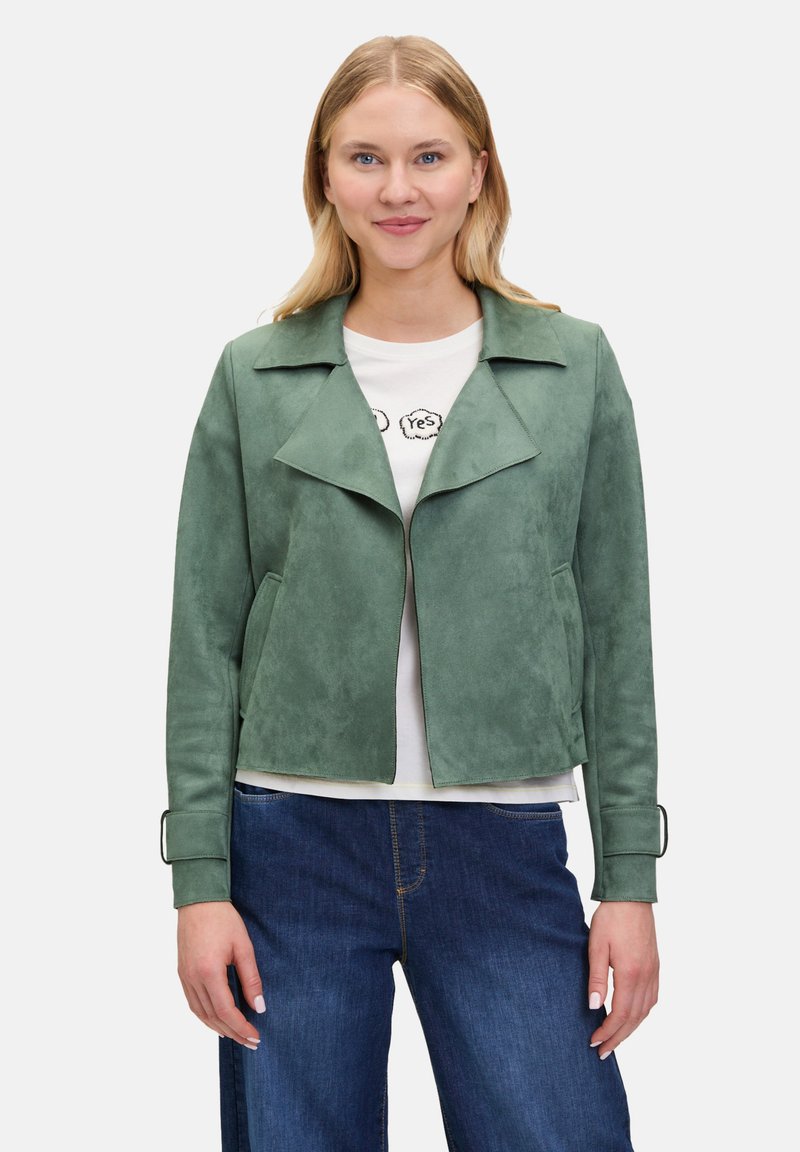 Veste en suédine verte avec col à revers, fermeture éclair et poches latérales. Dotée de poignets avec accents en boucle. Portée sur un T-shirt blanc et un jean bleu.