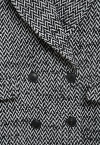 Tissu à motif pied-de-poule noir et blanc avec une texture douce, design croisé à double boutonnage, et deux boutons noirs sur le devant.