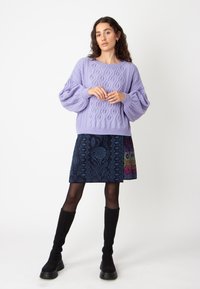 Pull en lilas tricoté avec des motifs en losange, associé à une jupe bleu foncé ornée de motifs floraux et géométriques dans des couleurs vives, bottes noires au genou.