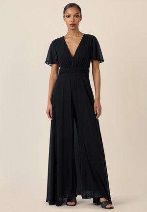 Kvinde står iført en sort, gulvlængde kjole med flagrende ærmer og dyb V-hals, kombineret med sorte åbne hæle, beige baggrund.