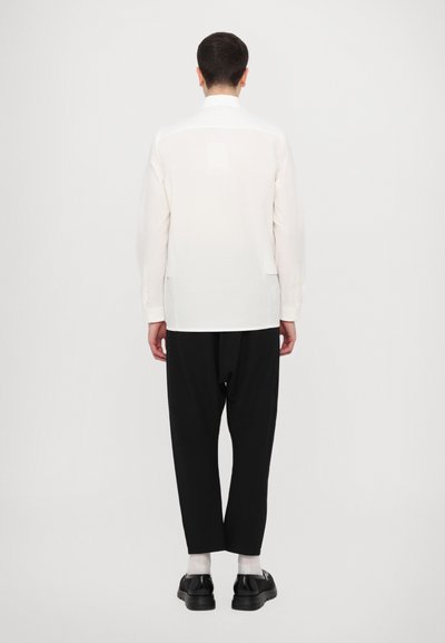 Camisa blanca de manga larga con dobladillo recto, combinada con pantalones negros de corte holgado y zapatos negros, vista desde la parte posterior contra un fondo liso.