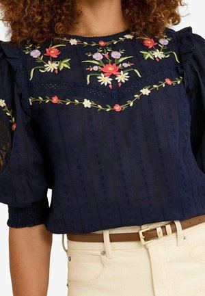 Kvinde iført en mørkeblå bluse med broderede farverige blomster og cremefarvede bukser med brun bælte.