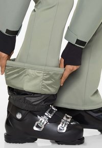Schneehosen mit einer hellgrünen Außenhülle, schwarzen elastischen Bündchen und einem glänzenden silbernen Reißverschluss. Schwarze Skischuhe mit metallischen Schnallen und mattem Finish.