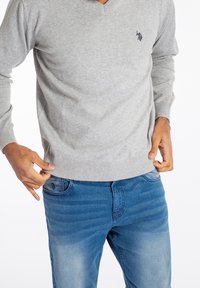 Man som bär en ljusgrå V-ringad tröja och blå jeans, justerar tröjans nederkant med båda händerna.