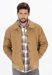 Veste en velours côtelé marron avec une fermeture à boutons-pression, tissu texturé, grand col et détail d'ancre brodé. Superposée sur une chemise en flanelle à carreaux.
