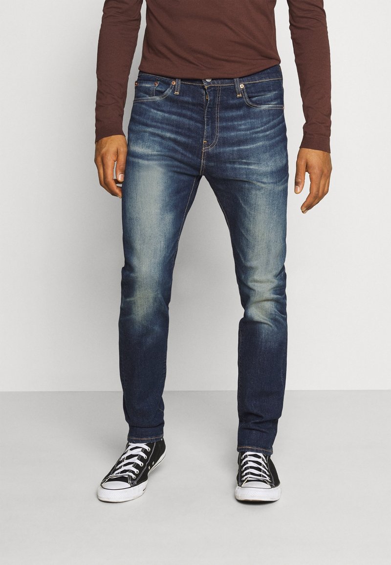 Homme portant un jean bleu délavé, des baskets noires et blanches, et une chemise marron à manches longues, debout devant un fond uni.