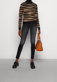 Pull à col roulé rayé noir et beige, jean gris slim, mocassins noirs épais et sac à main orange. Textures lisses, design moderne.