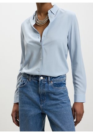 Person trägt ein hellblaues Hemd mit Knöpfen, geschichtete Perlenketten und hoch taillierte blaue Jeans, die Hände entspannt an den Seiten.