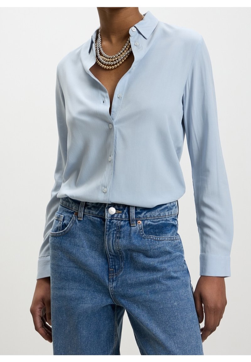 Personne portant une chemise bleu clair à boutons, des colliers de perles superposés et un jean bleu taille haute, les mains détendues le long du corps.
