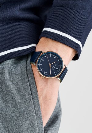 Navy blue watch with round face, gold accents, and a dark blue leather strap, worn on a wrist with a grey fabric sleeve.