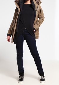 Parka verde oliva con cappuccio foderato in pelliccia, camicia nera lunga sotto, jeans skinny blu scuro e sneaker nere con dettagli in frange.