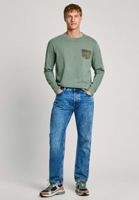 Camisa de algodón de manga larga en color verde oliva con un logo de bolsillo marrón, combinada con jeans de mezclilla azules y zapatillas multicolor.