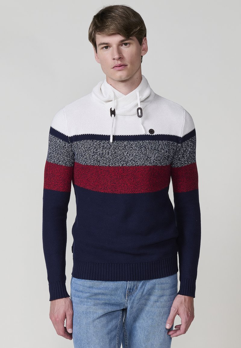 Joven vistiendo un suéter a rayas en colores navy, rojo y blanco, posando contra un fondo neutro, mostrando un estilo de moda relajado.