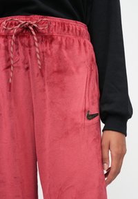 Osoba nosící červené sametové kalhoty Nike s nastavitelným pasem a černé tričko s dlouhým rukávem, ruka volně podél těla.