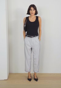 Haut sans manches noir, pantalon taille haute gris clair avec plis, talons noirs et un sac à main noir, sur un fond neutre.