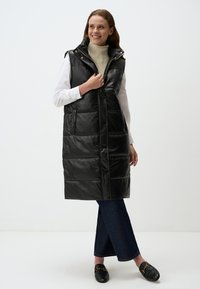 Gilet matelassé sans manches noir avec une finition brillante, des boutons sur le devant, des poches latérales et un col haut ; porté sur un col roulé crème et un jean bleu.