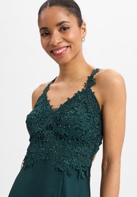 Emeraldgroene jurk met ingewikkelde bloemenkantdetails bij het lijfje, een V-halslijn en dunne bandjes. Soepele satijnen rok.