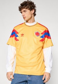 Maglia da calcio gialla con maniche a righe blu e rosse, con colletto rotondo e logo Adidas sul davanti. Dettaglio del tessuto testurizzato visibile.