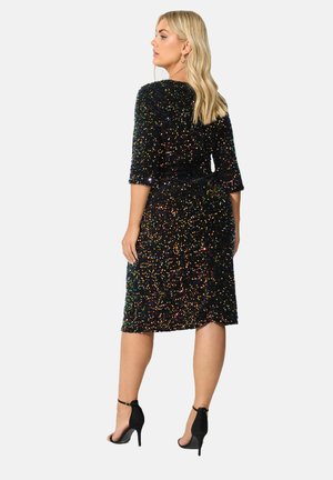 Abito nero con paillettes, maniche a tre quarti, lunghezza ginocchio. Presenta paillettes multicolori, una vita aderente e una leggera lucentezza. Indossato con tacchi neri.
