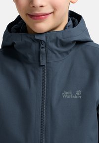 Giacca a vento blu scuro con cappuccio, dotata di zip frontale completa, caratterizzata da un tessuto strutturato e un logo sul petto. Finitura liscia con cappuccio regolabile.