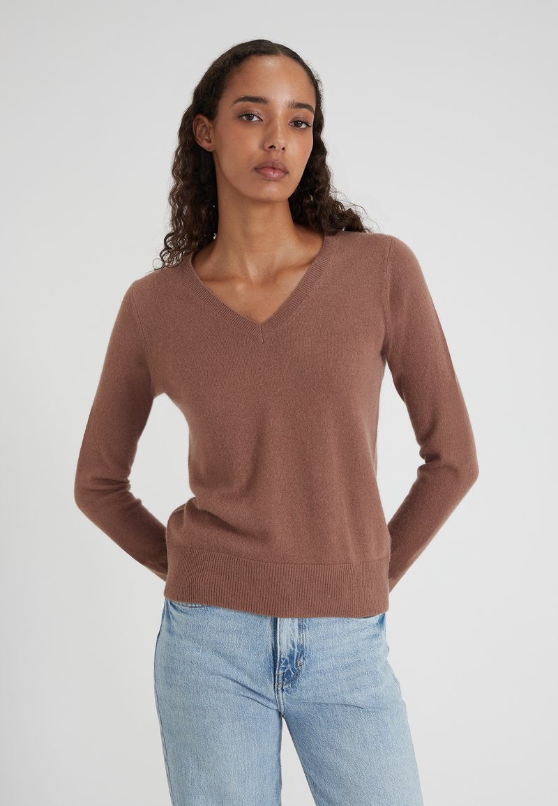 Donna con lunghi capelli ricci che indossa un maglione brown a V e jeans azzurri, in piedi con le mani dietro la schiena su uno sfondo neutro.