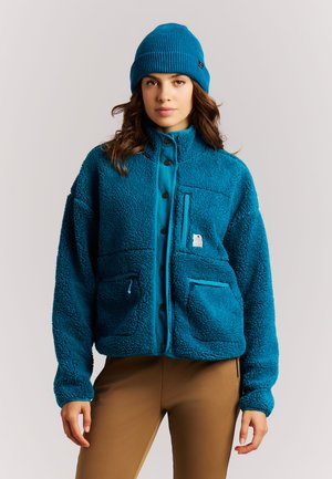 Woman wearing a blue fleece jacket with buttoned front, matching blue knit beanie, and brown pants, standing against a plain background.