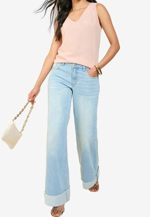 Femme portant un débardeur rose clair et un jean large bleu clair, tenant un petit sac à main beige avec une bandoulière chaîne dorée.