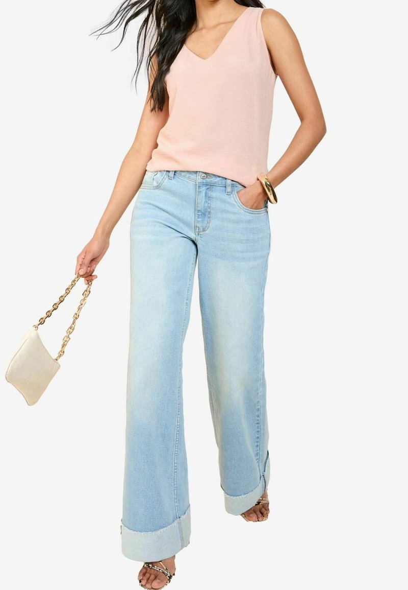 Femme portant un débardeur rose clair et un jean large bleu clair, tenant un petit sac à main beige avec une bandoulière chaîne dorée.