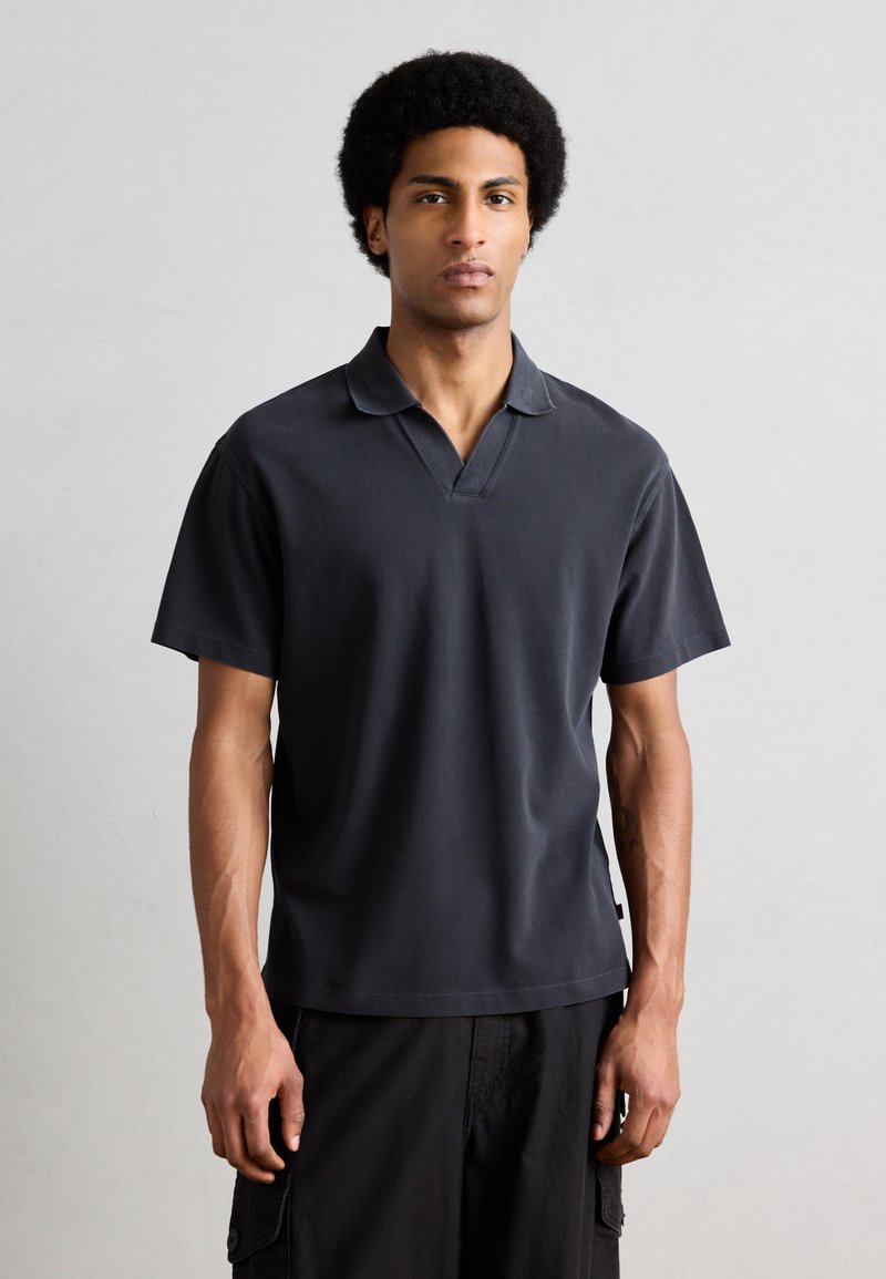 Νεαρός άνδρας με κοντό αφρο-κράτημα, φοράει σκούρο γκρι πόλο με κοντά μανίκια και μαύρο παντελόνι cargo, στέκεται μπροστά σε απλό ανοιχτόχρωμο φόντο.