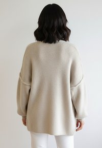 Beżowy, dziergany sweter o luźnym kroju, z prążkowanym dekoltem i szerokimi bufkami na rękawach. Materiał jest miękki, a długość sięga do połowy tułowia.