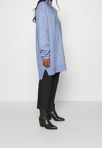 Blå stickad tröja med hög krage, sidofickor och långa ärmar, i kombination med svarta jeans och svarta spetsiga ankelskor.