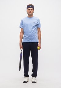 Νέος άνδρας με ανοιχτό μπλε μπλουζάκι, σκούρα μπλε παντελόνια και κορδέλα στο κεφάλι, κρατώντας ρακέτα και μπάλα τένις, στέκεται μπροστά σε λευκό καθαρό φόντο.