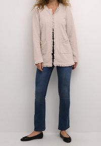 Jasnobeżowa, teksturowana kurtka z falbaną, dwie przednie kieszenie, zestawiona z ciemnoniebieskimi dżinsami i czarnymi balerinami.