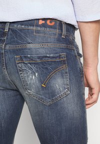 Dżinsy z denimu w ciemnym niebieskim kolorze o przetartym wyglądzie, z wyraźnym pomarańczowym logo nad tylną kieszenią oraz kontrastującym żółtym przeszyciem na kieszeniach.